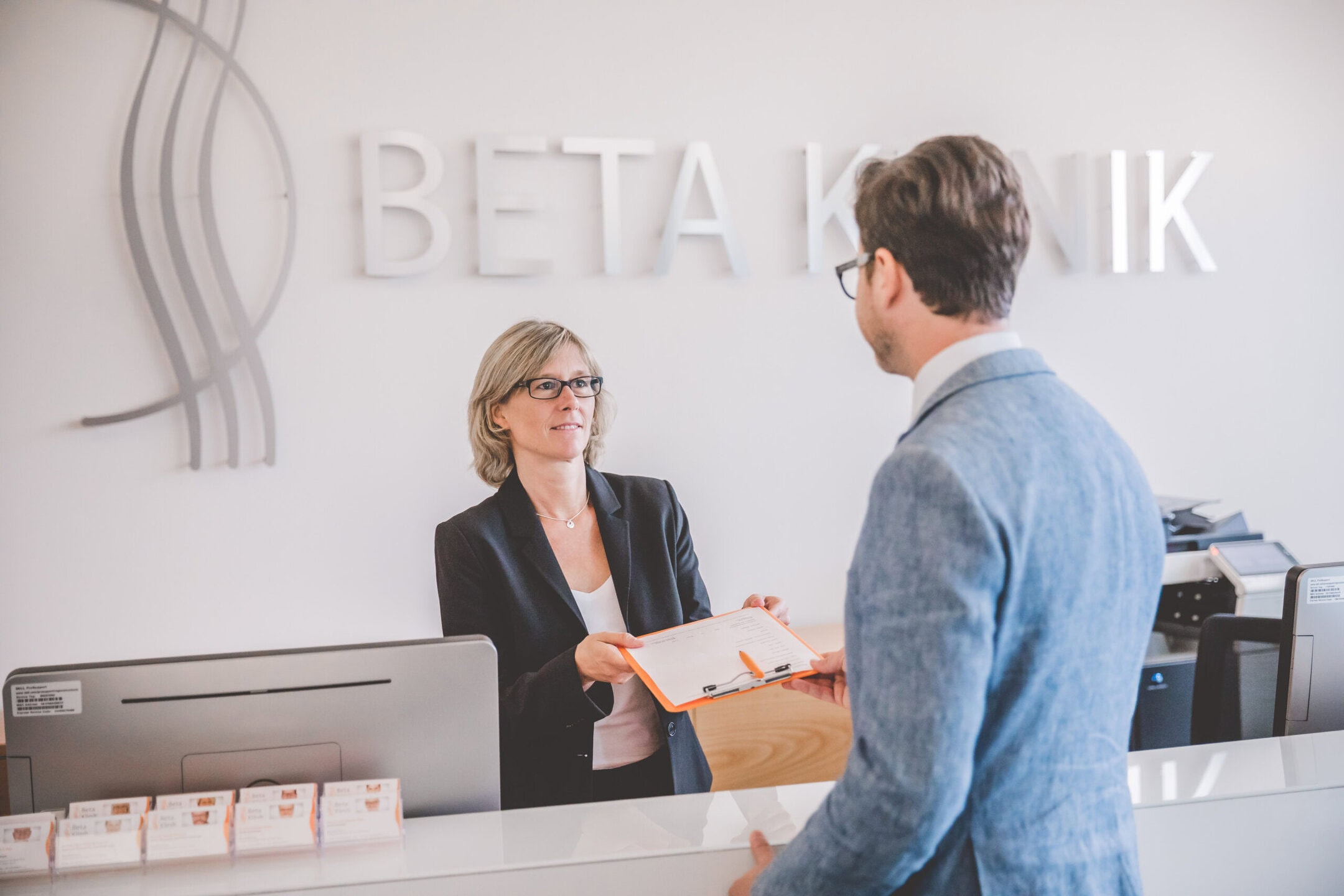 Empfangs Mitarbeiterin händigt einem Patienten ein Klemmbrett aus.