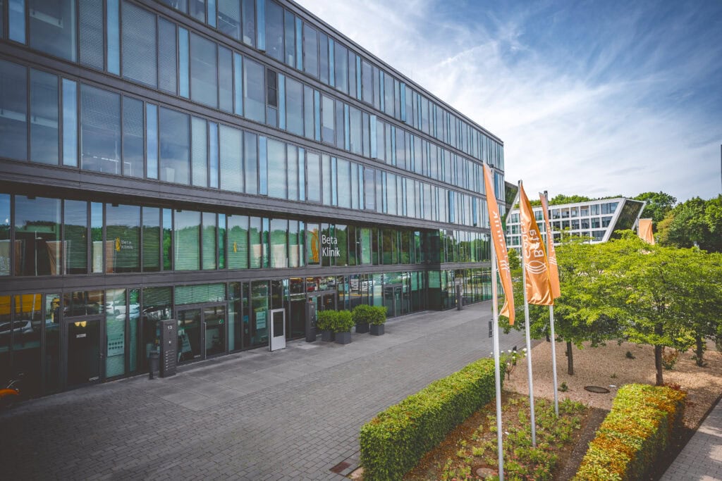 Moderne Glasfassade eines Gebäudes mit drei orangefarbenen Fahnen und grünen Bäumen im Vordergrund.