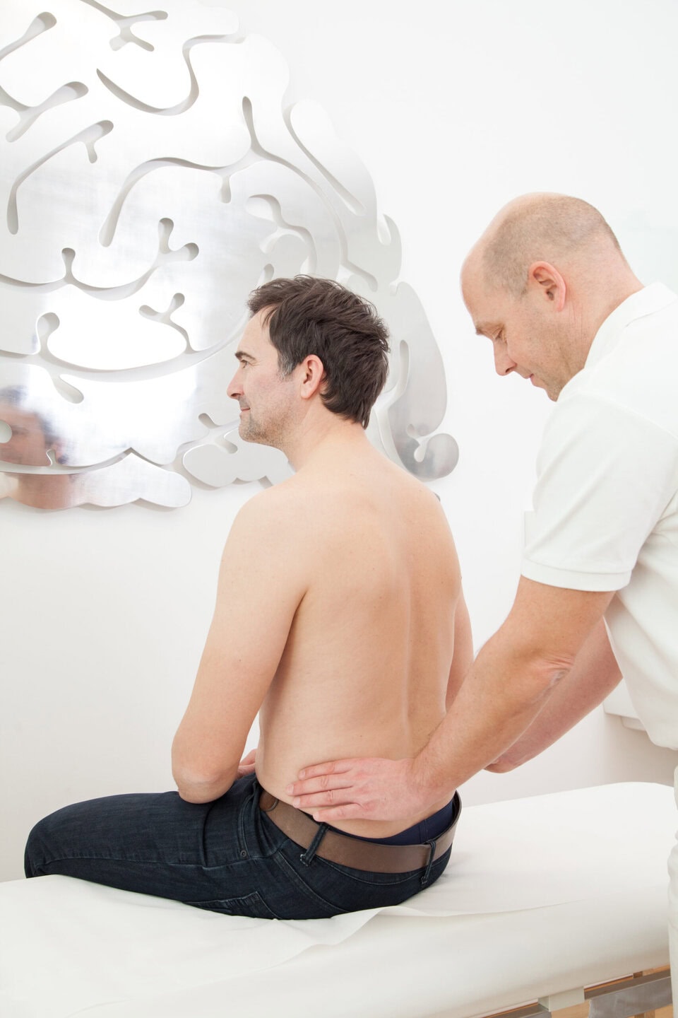 Ein Mann ohne Hemd sitzt, während ein anderer Mann seinen unteren Rücken untersucht.