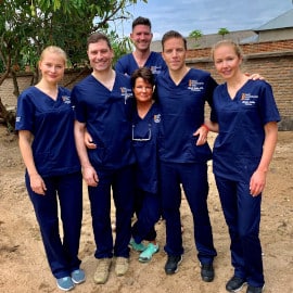 Sechs Personen in blauen medizinischen Uniformen stehen draußen auf sandigem Boden und lächeln in die Kamera.