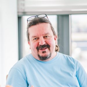 Ein lächelnder Mann mit Brille auf dem Kopf trägt ein hellblaues Shirt und sitzt vor einem Fenster.