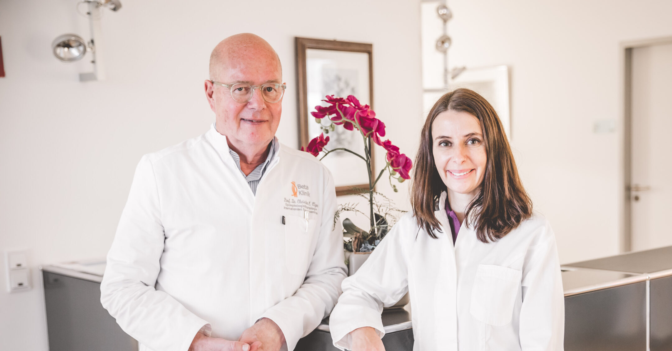 Zwei Ärzte in weißen Kitteln stehen lächelnd vor einer Orchidee in einer Praxis.
