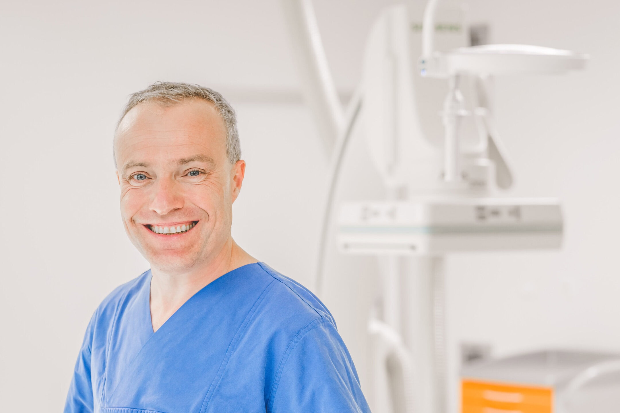 Ein lächelnder Mann in blauer OP-Kleidung steht vor medizinischen Geräten.