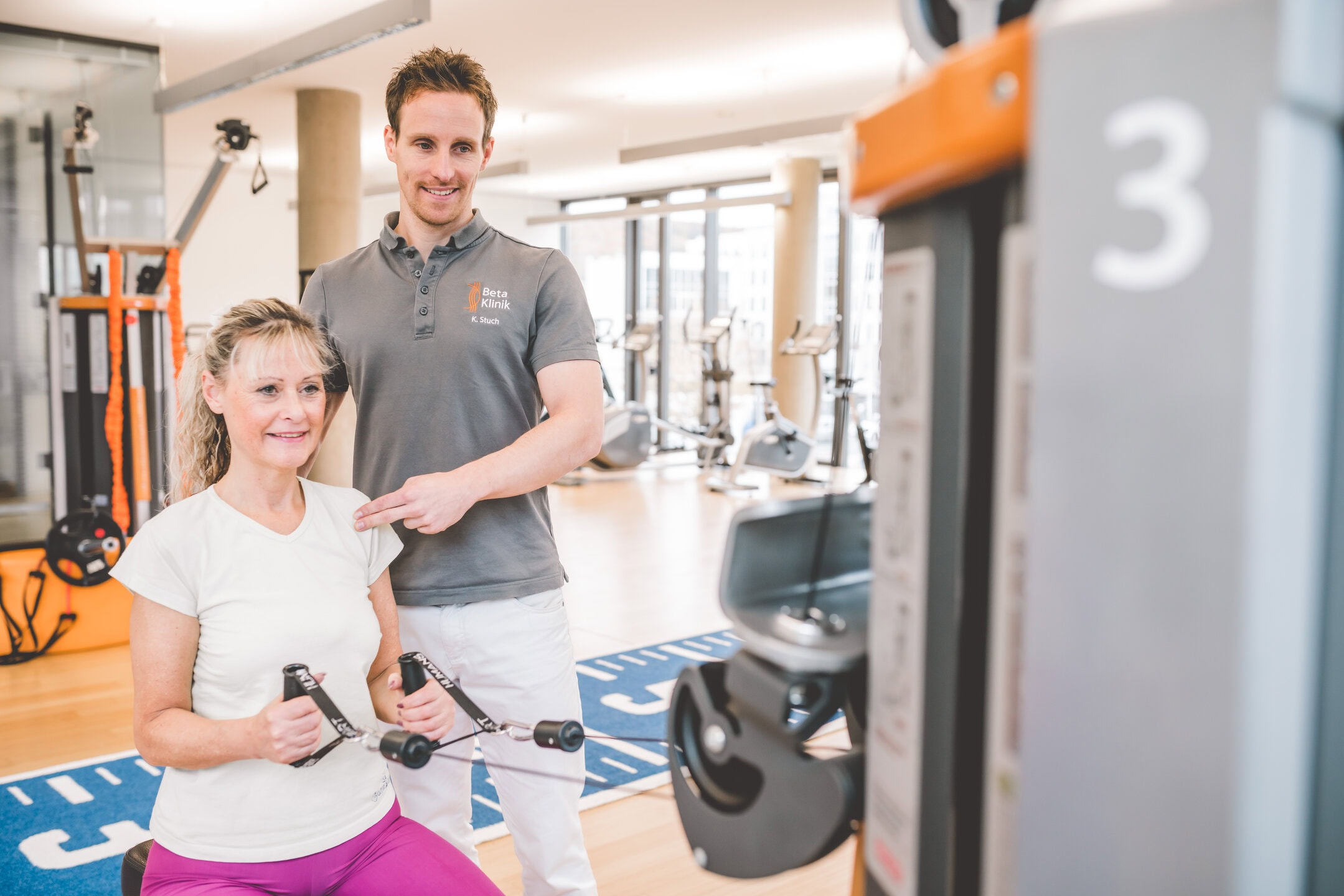 Eine Frau trainiert an einem Fitnessgerät, während ein Trainer neben ihr steht und sie anleitet.