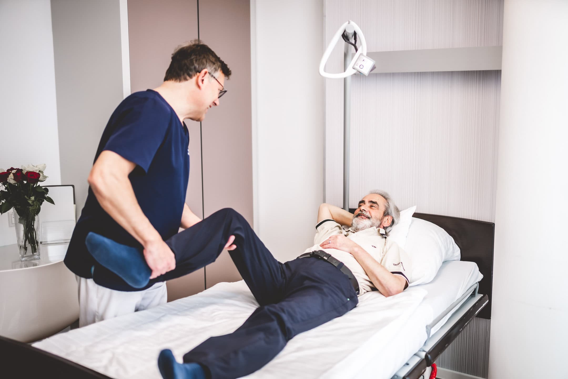 Ein Mann liegt lächelnd auf einem Bett, während ein Pfleger sein Bein anhebt.