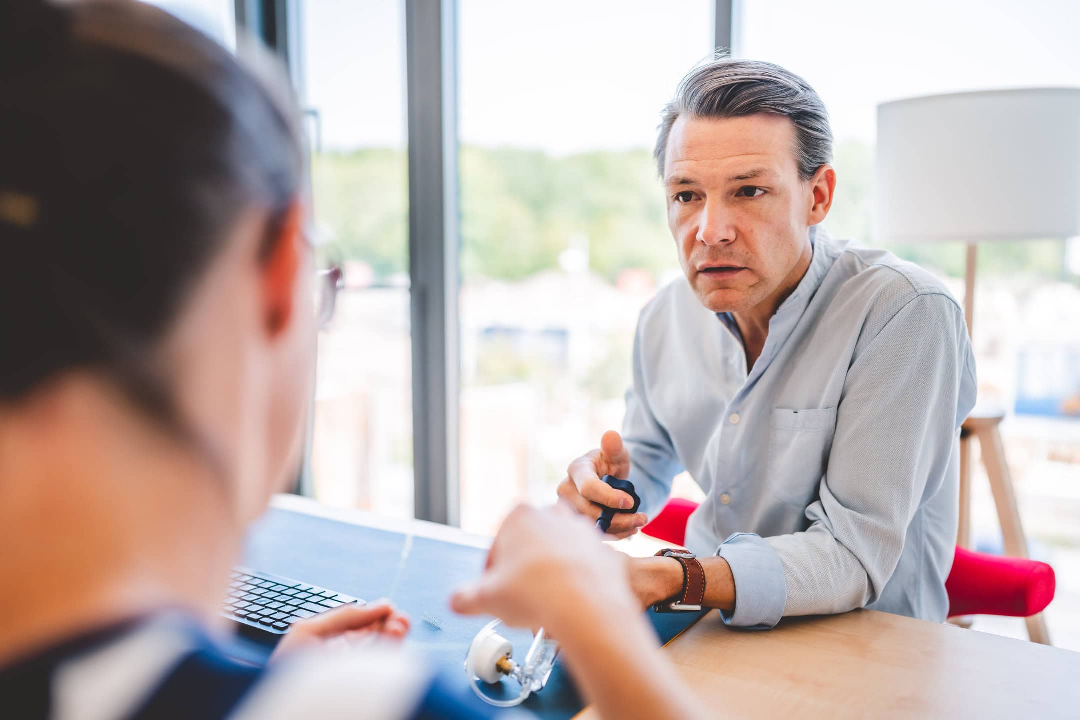Ein Mann im Gespräch mit einer Frau, beide sitzen an einem Tisch vor einem großen Fenster.