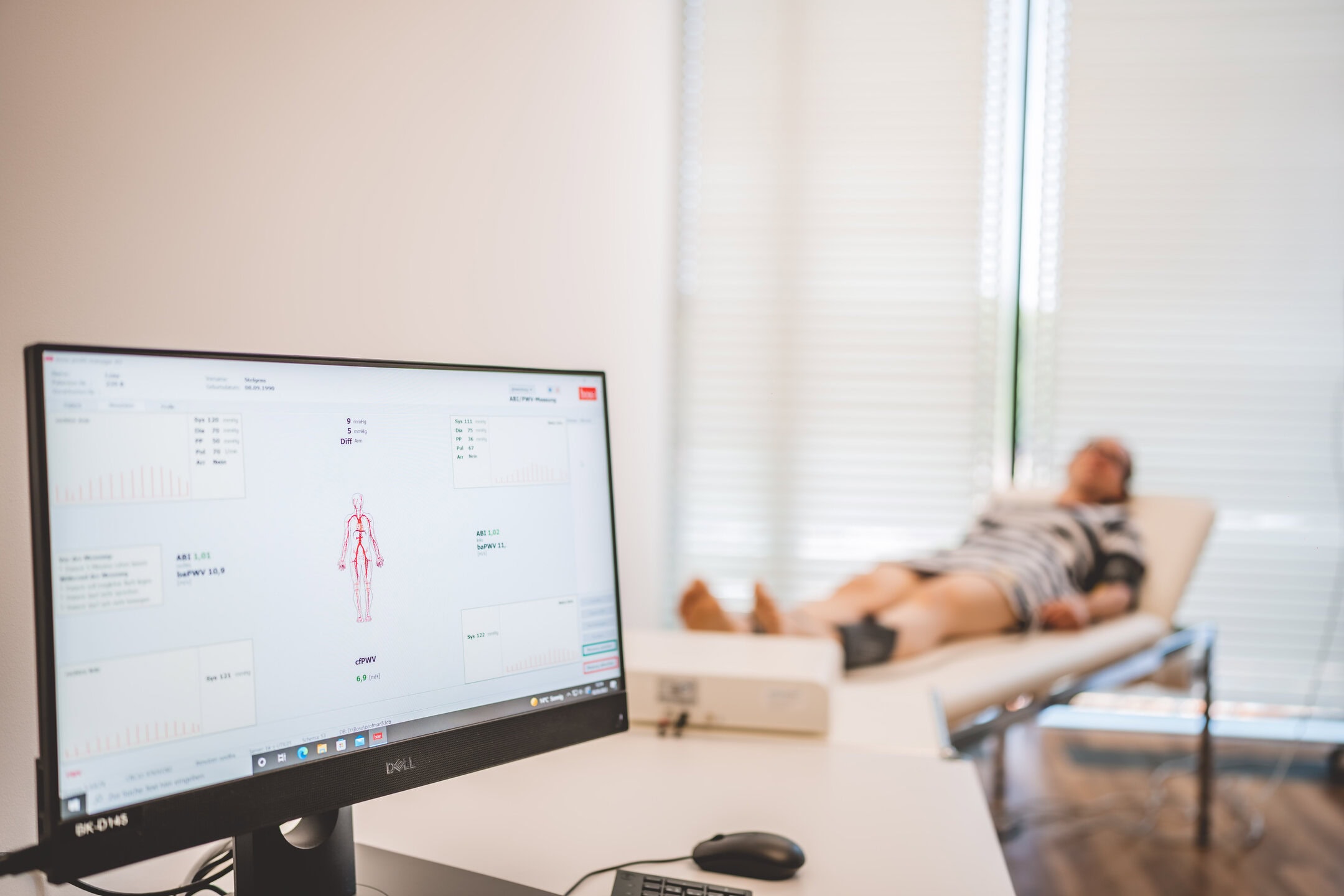 Ein Computerbildschirm zeigt medizinische Daten, während eine Person auf einer Liege im Hintergrund liegt.