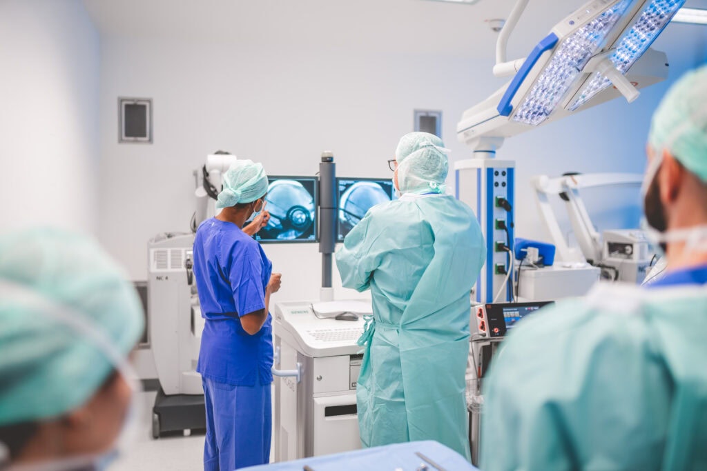 Ärzte in OP-Kleidung betrachten Röntgenbilder auf Monitoren in einem hellen Operationssaal.