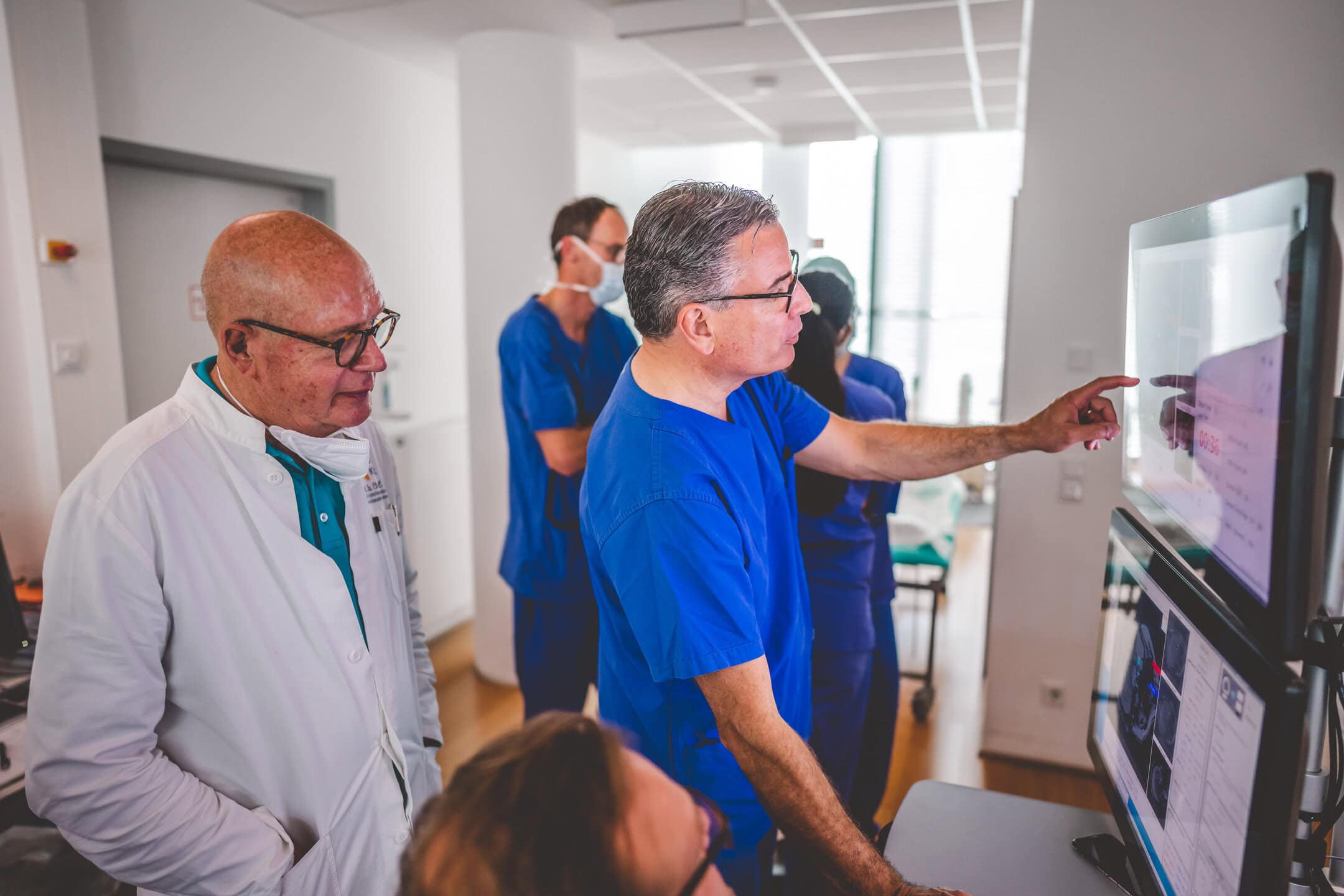 Ärzte in blauen Kitteln diskutieren vor Monitoren in einem hellen Raum.