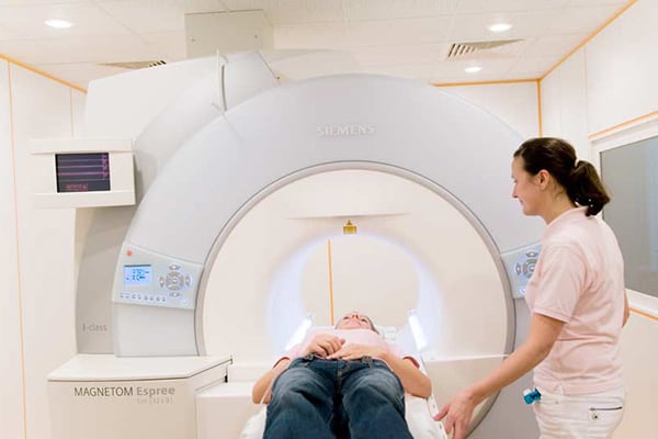Eine Person liegt in einem MRT-Gerät, betreut von einer lächelnden Frau in einem weißen Raum.