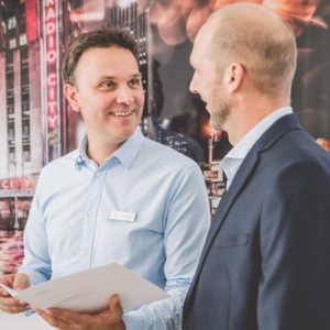 Zwei Männer im Gespräch vor einem Hintergrund mit Neonlichtern; einer hält Papiere.