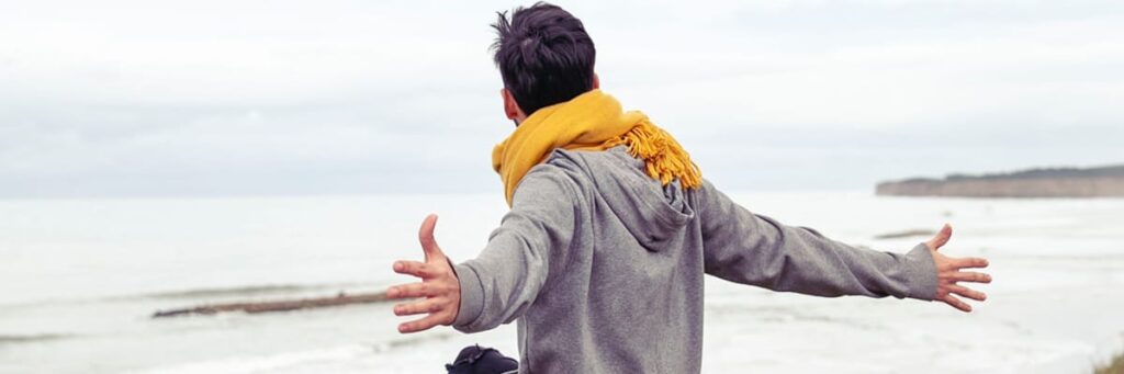 Person mit gelbem Schal breitet die Arme aus, steht am Meer unter bewölktem Himmel.