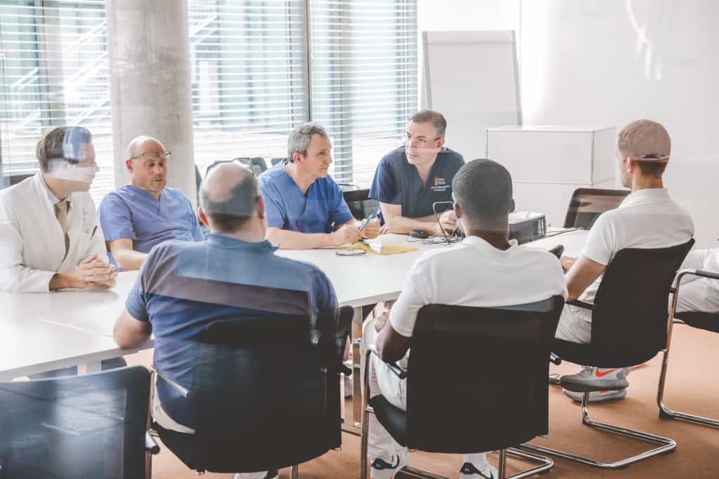 Sieben Personen in medizinischer Kleidung sitzen um einen Konferenztisch in einem hellen Raum.