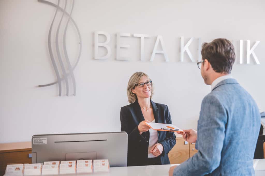 Patient wird am Empfang der Beta Klinik freundlich von der Mitarbeiterin begrüßt und bekommt einen Anmeldebogen überreicht