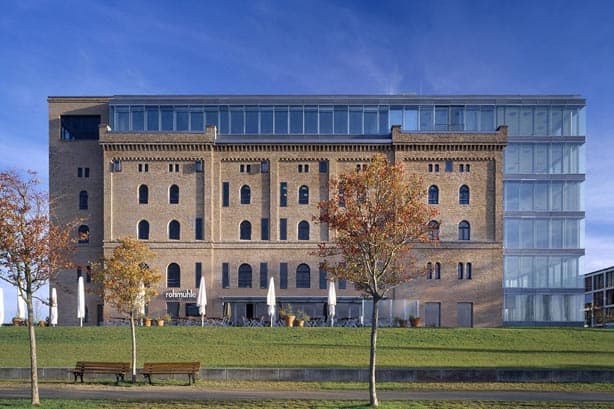 Ein historisches Backsteingebäude mit moderner Glasfassade, Bäume und Bänke im Vordergrund.