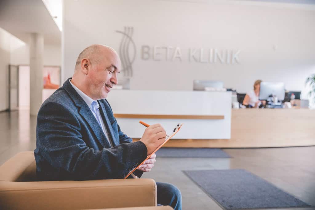 Ein Mann sitzt in einer Klinik und füllt ein Formular aus, während im Hintergrund eine Rezeptionistin arbeitet.