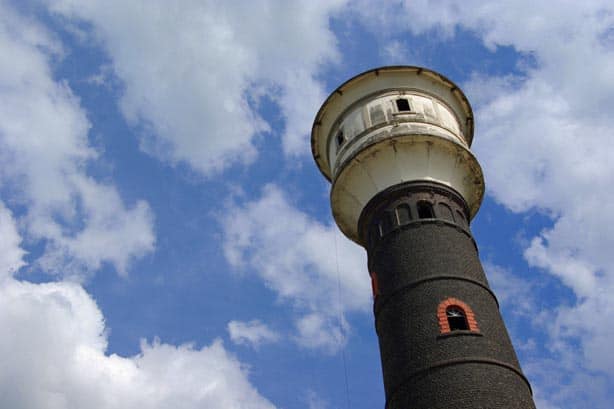 Ein alter Wasserturm mit dunklen Ziegeln vor blauem Himmel mit weißen Wolken.
