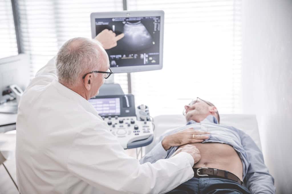 Ein Arzt führt eine Ultraschalluntersuchung am Bauch eines liegenden Patienten durch.