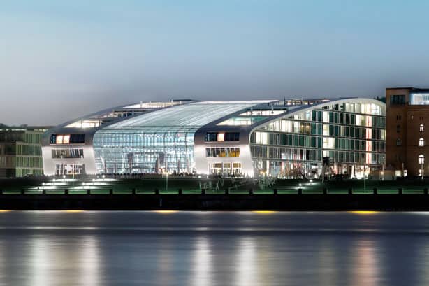 Ein modernes, beleuchtetes Gebäude mit Glasfassade am Flussufer bei Abenddämmerung.