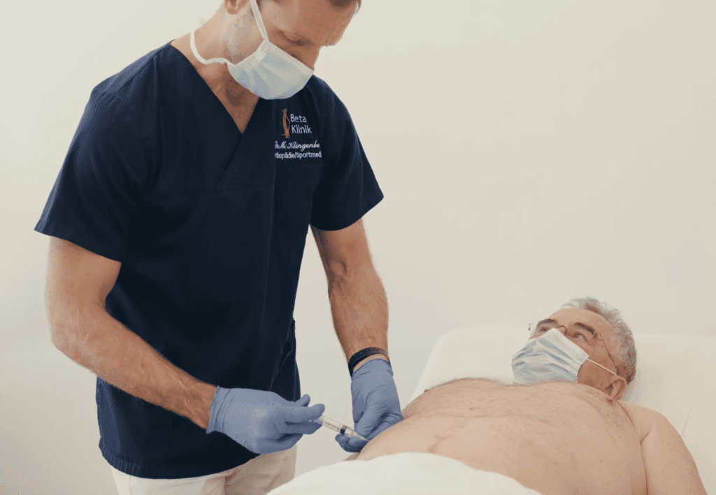 Ein Arzt in blauer Kleidung injiziert einem liegenden Mann eine Spritze in den Bauch. Beide tragen Masken.