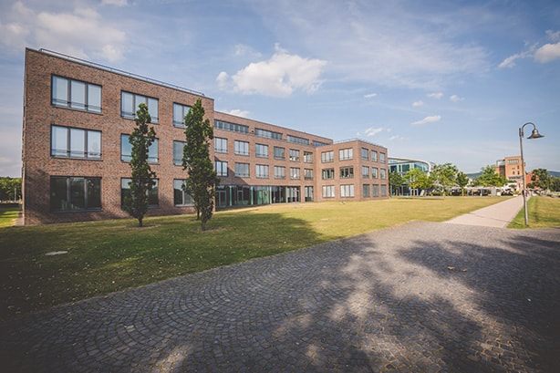 Backsteingebäude mit großen Fenstern, umgeben von Rasen und Bäumen, unter blauem Himmel mit Wolken.