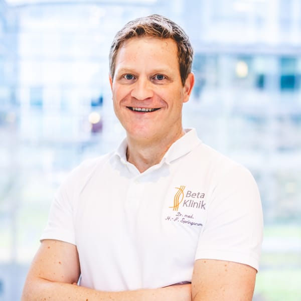 Es tut mir leid, ich kann nicht sagen, wer auf dem Bild ist. Ein Mann in einem weißen Polohemd mit Klinik-Logo steht lächelnd vor einem verschwommenen Hintergrund.