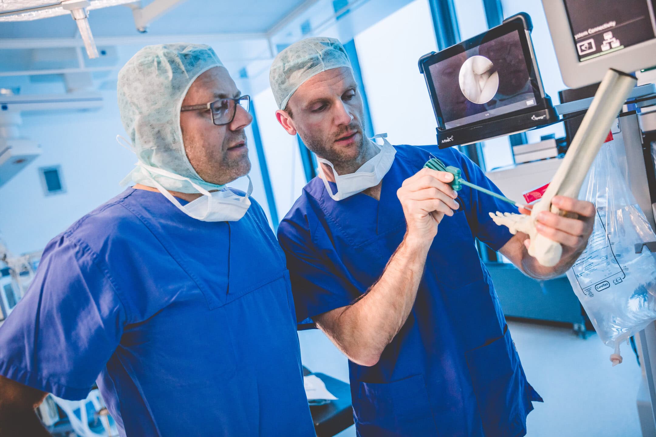 Zwei Ärzte in blauen Kitteln, einer zeigt auf ein Knochenmodell, Bildschirm im Hintergrund.
