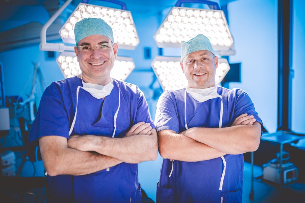 Zwei Norologen stehen im OP mit OP-Klamotten und schauen in die Kamera.