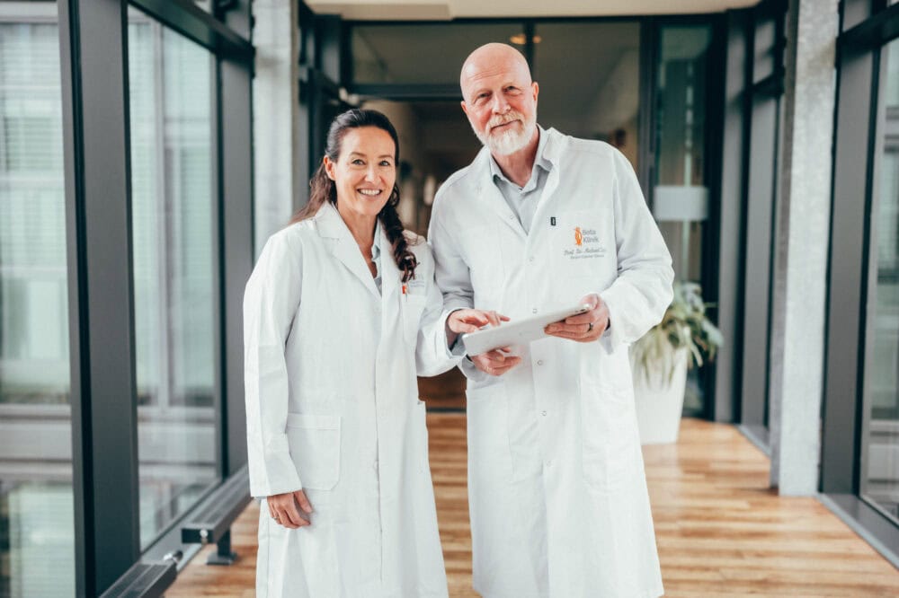 Zwei Ärzte in weißen Kitteln stehen lächelnd in einem hellen Flur mit Holzfußboden.