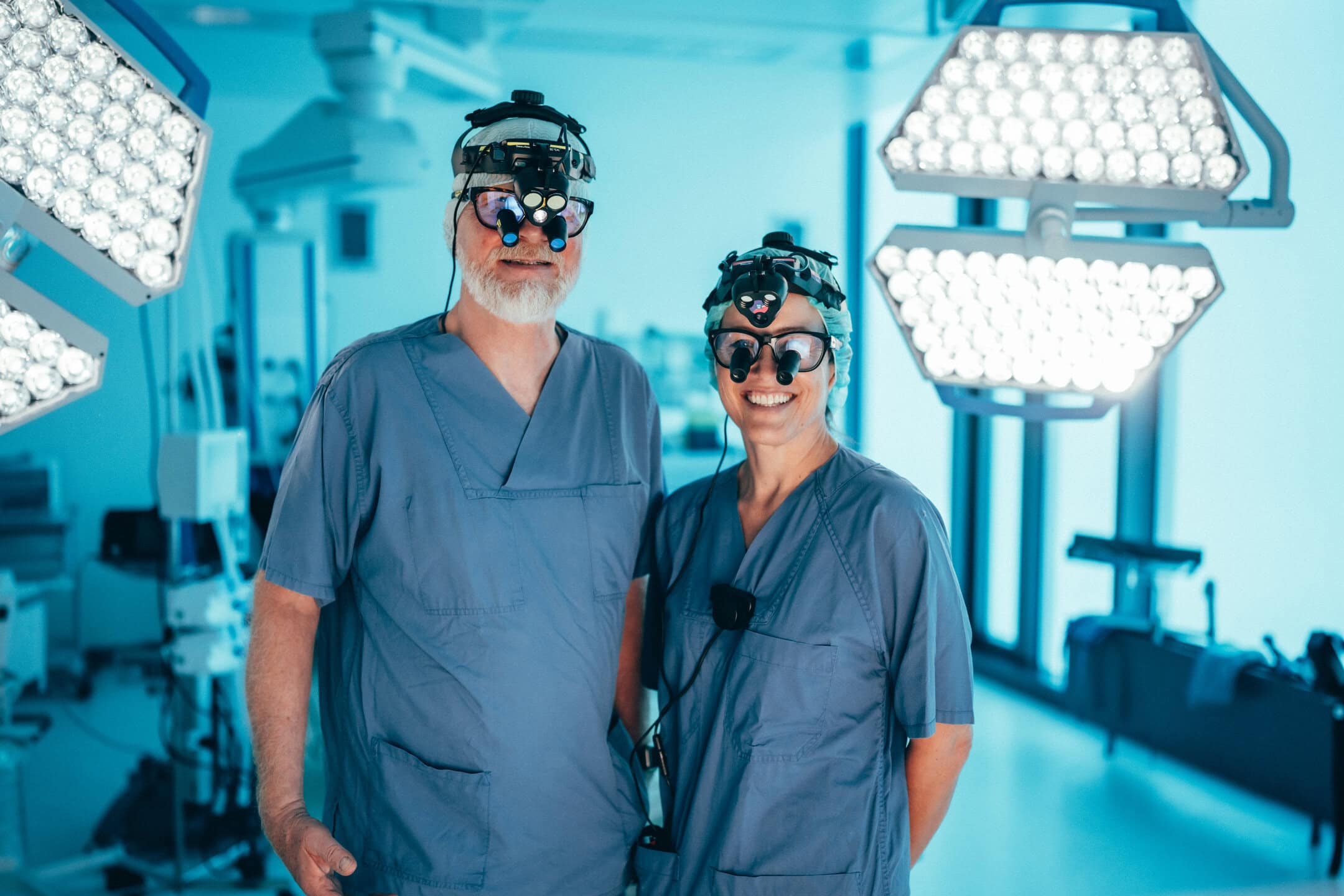 Zwei Chirurgen in blauen Kitteln mit Lupenbrillen, umgeben von OP-Leuchten in einem hellen Raum.