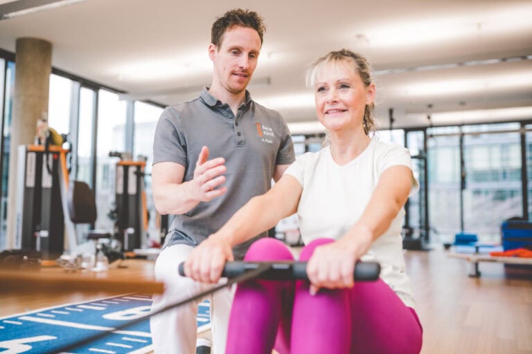 Patientin trainiert am Rudergerät und wird dabei vom Physiotherapeuten begleitet