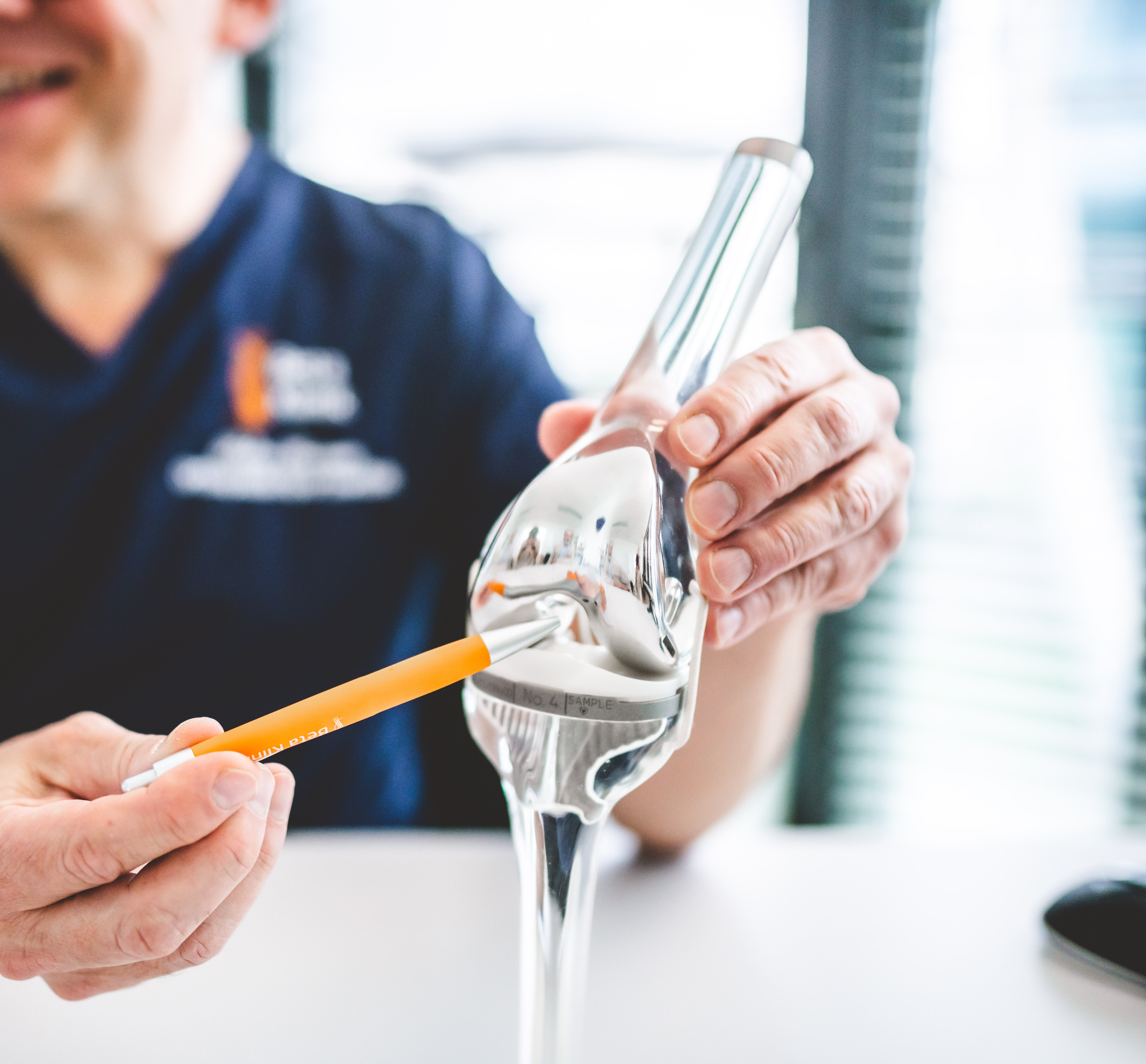 Eine Person hält ein silbernes Kniegelenkmodell und zeigt mit einem Stift darauf.