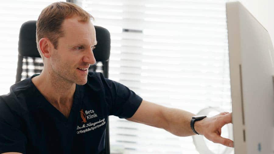 Ein Mann in dunkler Kleidung zeigt auf einen Computerbildschirm in einem hellen Büro.