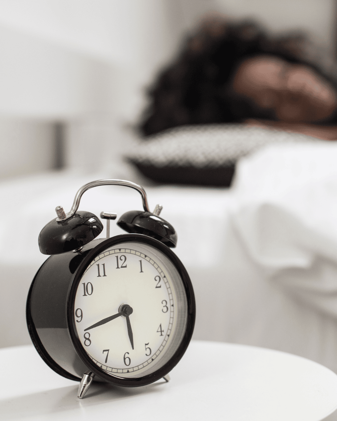 Schwarzer Wecker zeigt 7:00 Uhr auf weißem Tisch, unscharfe Person im Bett, symbolisiert erholsamen Schlaf und zirkadianen Rhythmus.