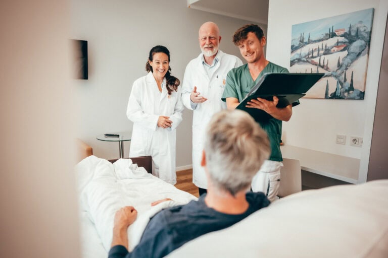 Prof. Sabel und Prof. Rapp gemeinsam mit einem Pfleger am Krankenbett in warmherziger, wertschätzender Atmosphäre